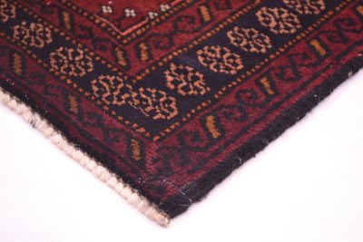 A close-up corner view of a patterned Maroon Afghan Belouch mat on a white background, showing a dark navy blue border with repeating beige floral motifs, a burgundy-red border, and a cream-coloured twisted fringe.