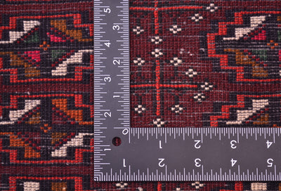 Close-up of the hand-knotted weave of a patterned Afghan Belouch mat with a dark maroon and black geometric design, shown alongside a metal ruler to show scale of the knotting.