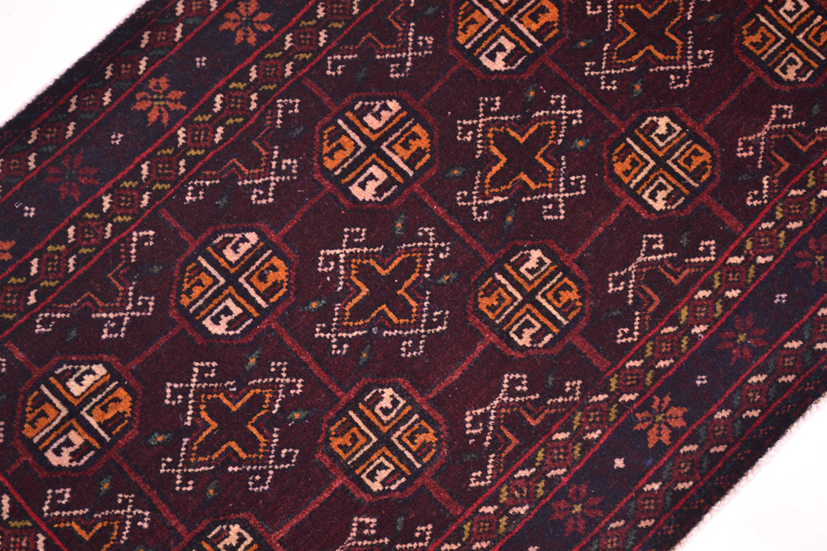 A close-up of a patterned Bokhara design Belouch mat with a dark burgundy background and geometric motifs in orange, beige, and white.