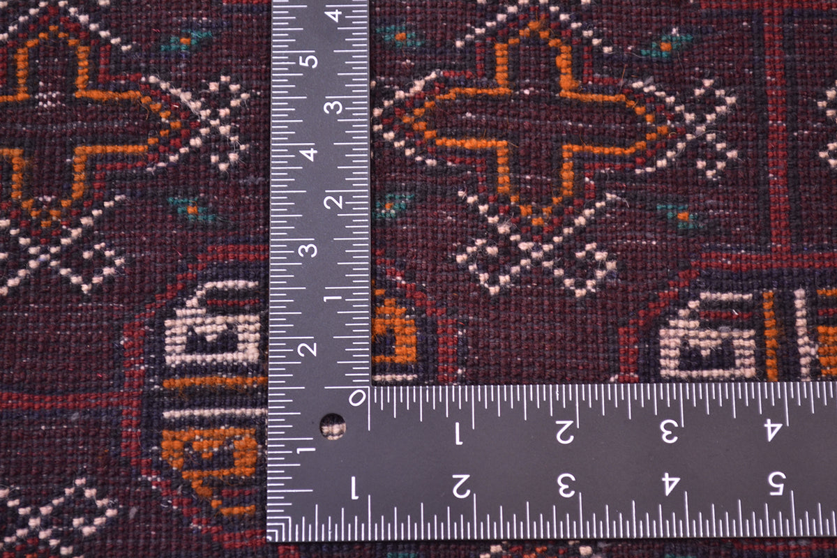 A close-up of the hand-knotted back of a patterned Bokhara design Belouch mat with a dark reddish-brown base and geometric motifs in orange, cream, and teal, shown with a metal ruler to show scale of the knot density.