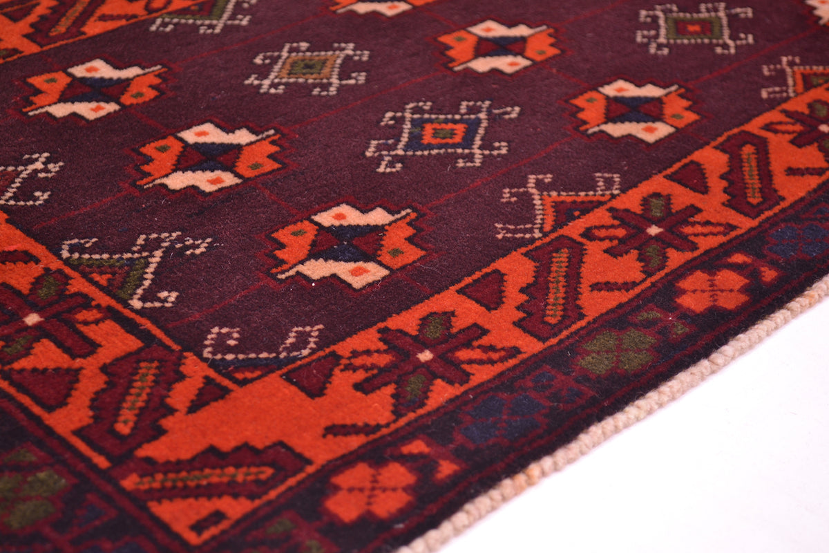 A close-up of a patterned afghan rug showing a dark brown/plum-purple field with orange, red, beige, and green geometric motifs and borders.