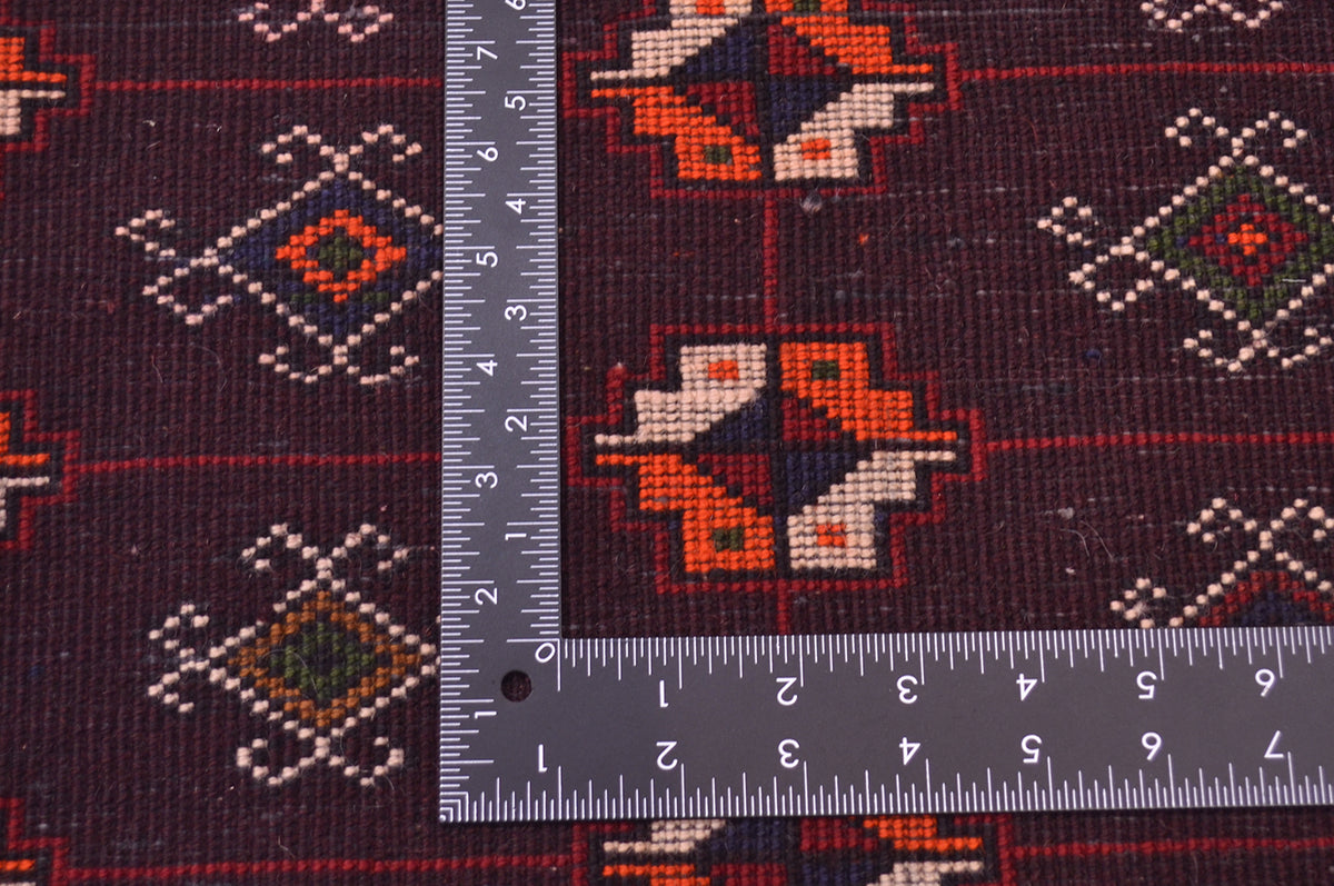 A close-up of a dark patterned rug with geometric motifs in red, orange, cream, and green, shown with a metal L-square ruler to indicate scale.