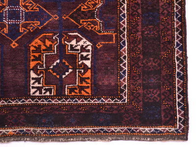 A close-up of an old tribal-style Belouch rug corner, showcasing intricate geometric and tribal patterns in deep burgundy, orange, navy blue, and cream.