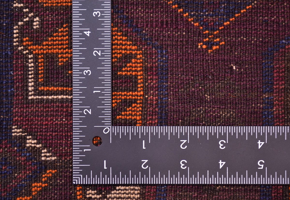 Close-up of the hand-knotted weave of a patterned Old Baluchi Afghan rug with a dark purple-brown base and orange, beige, and blue geometric motifs, shown with a metal ruler to show scale of the knotting.
