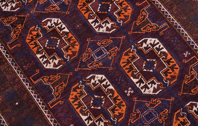 A close-up of a patterned old Afghan Belouch rug featuring geometric and tribal motifs in dark brown, rust orange, cream, and navy blue.