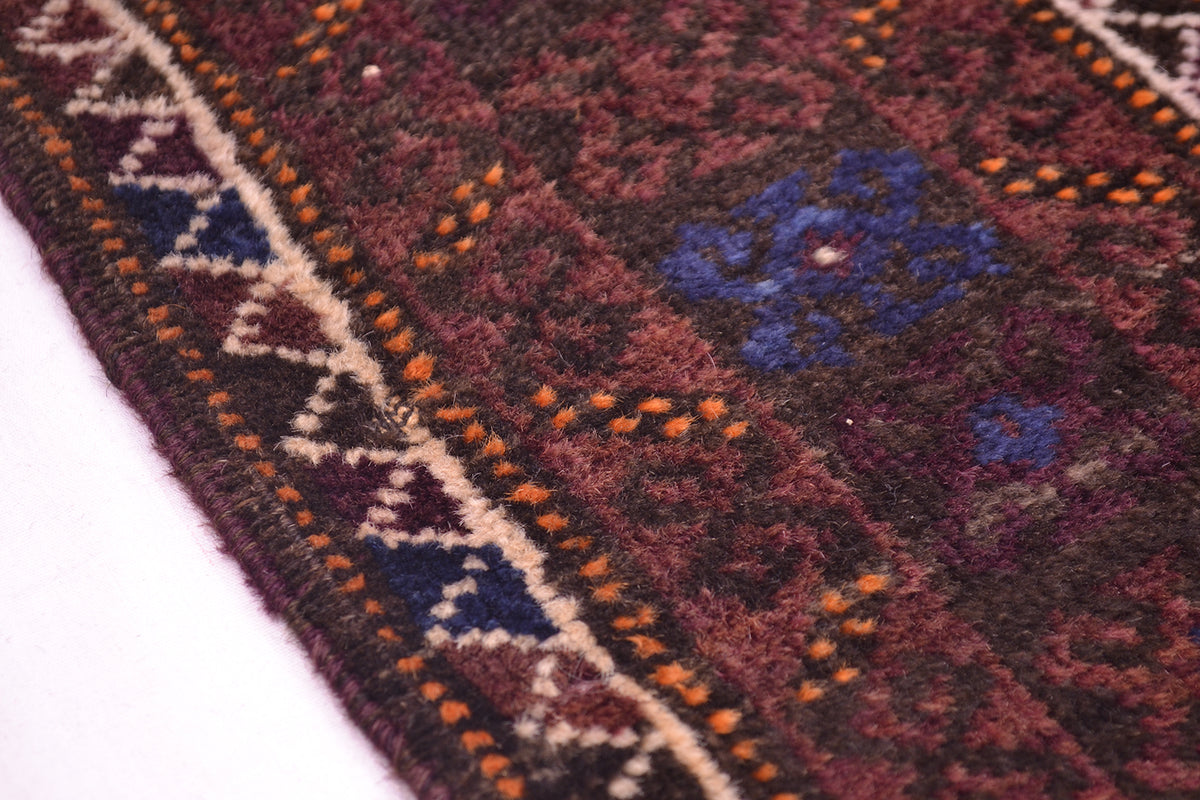 A close-up of a patterned old Afghan Baluch wool rug showing a dark brown and burgundy field with blue floral motifs, orange dotted accents, and a cream geometric border.