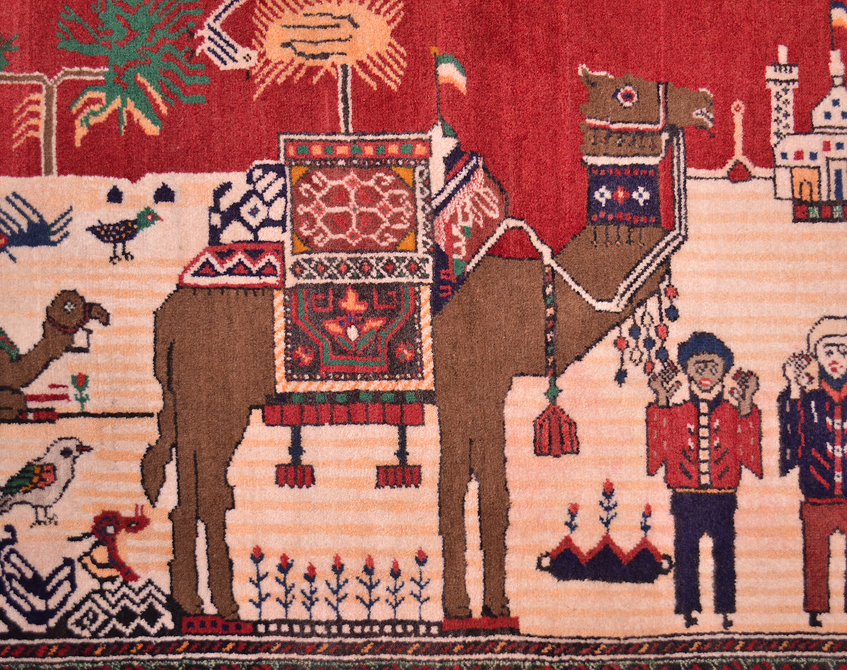 Close-up of a handwoven Afghan Pictorial Belouch rug featuring a camel with ornate red and patterned saddle, stylised figures, birds, and a red sky background.
