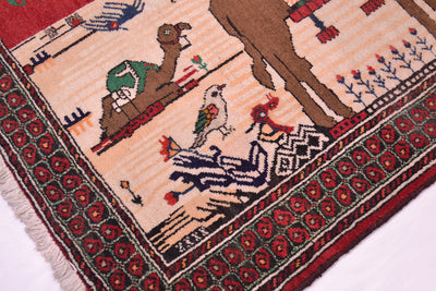 A close-up of a patterned Pictorial Belouch rug showing a camel, birds, and geometric floral motifs in red, beige, brown, black, and green.