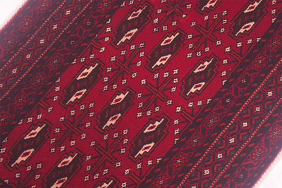 A close-up of a Afghan handmade red Belouch mat with repeating geometric gul motifs in dark brown, beige, and white.