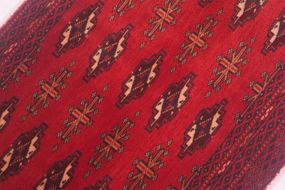 A close-up of a red patterned handmade Afghan Belouch mat, featuring repeating geometric motifs in dark brown, beige, and orange-brown.