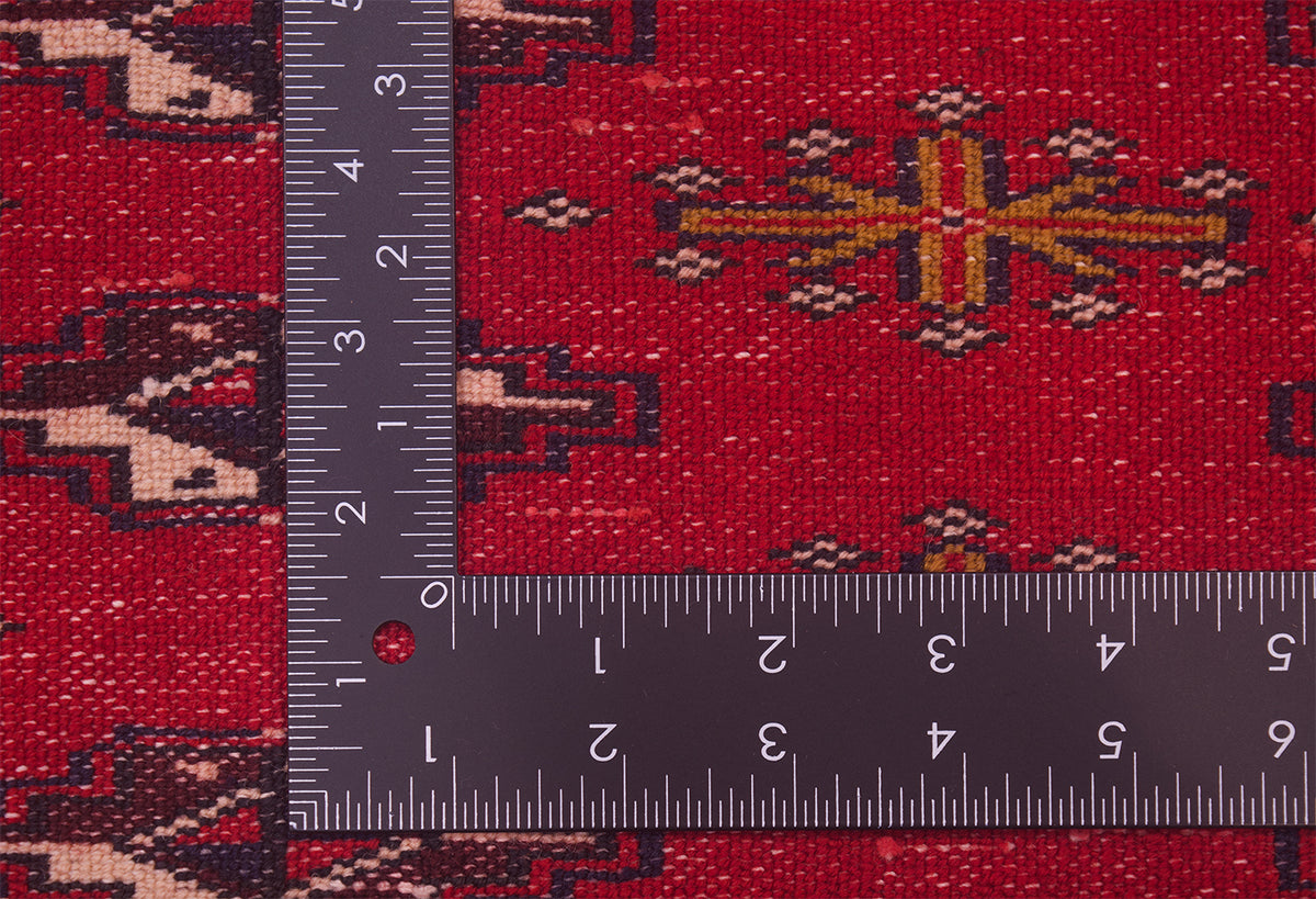 A close-up of the hand-knotted  back of a handmade red Afghan Belouch mat with geometric motifs, shown with a dark metal L-square ruler to indicate scale of the knot density.