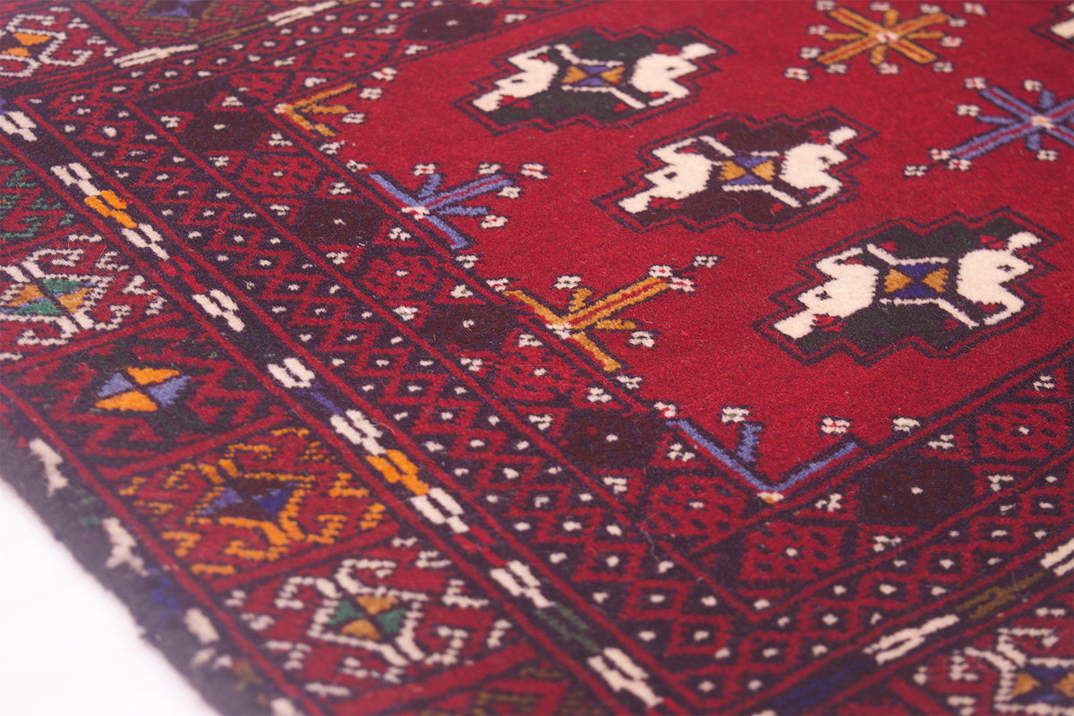 A close-up of a red patterned Afghan Belouch mat with geometric borders and stylised motifs in cream, black, blue, yellow, and white.