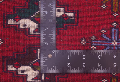 Close-up of the hand-knotted weave of a colourful red Afghan Belouch mat or small rug with a dark metal L-shaped ruler placed over it to show scale of the knotting, highlighting the detailed woven texture and measurements.