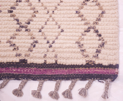 Close-up of a woven runner with a cream base, charcoal geometric patterning, a purple and dark stripe kilim border, and tasseled fringes along the bottom edge.