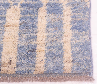 A close-up of a textured, shaggy woven rug with vertical stripes in light blue and cream, featuring a fringed edge.