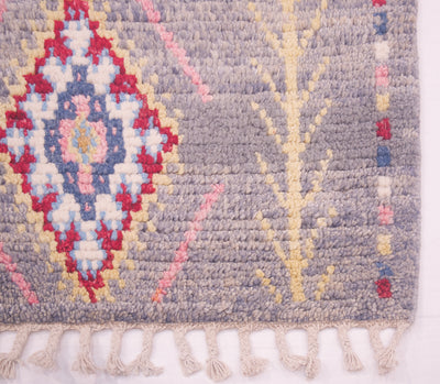 A close-up of a handwoven Moroccan-style Afghan rug corner showing a grey base with a multicolour diamond medallion pattern in red, cream, blue, and pink, plus a cream branch motif and fringe tassels along the bottom edge.