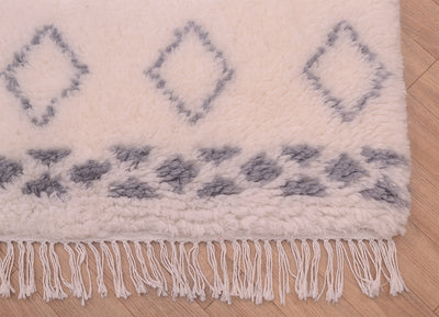 A close-up of a plush cream-colored shaggy pile Berber rug with a grey geometric diamond pattern and fringe tassels, laid on a light wood floor.