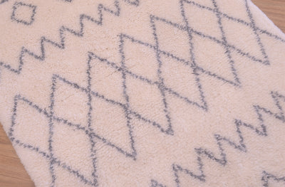 A close-up of a plush cream-colored Berber rug with a grey geometric diamond and zigzag pattern, laid on a light wood floor.