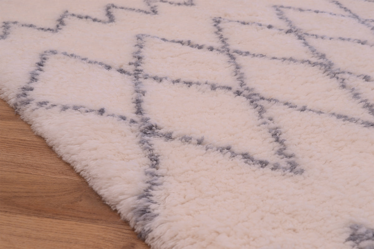 A close-up of a plush cream-colored shaggy berber rug with a grey geometric diamond pattern, shown partially folded over to reveal a light wood floor underneath.