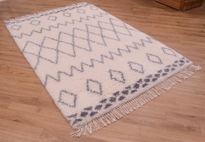 A rectangular shaggy pile Berber rugwith a cream base and grey geometric diamond and zigzag patterns, featuring fringe on two short ends, displayed on a light brown wooden floor.