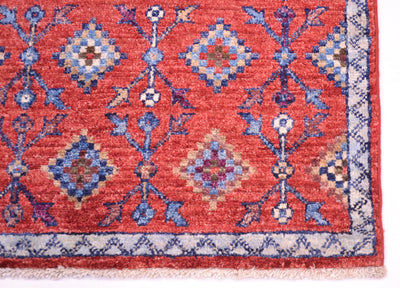 A close-up of a traditional patterned Chobi Afghan rug/mat corner, featuring a coral-red field with blue, beige, and purple geometric medallions and a cream border.