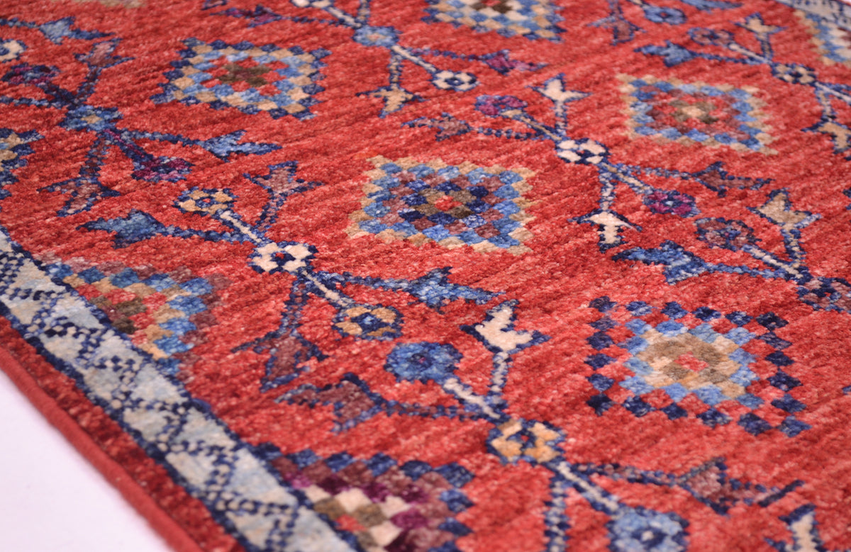 A close-up of a red patterned Afghan Chobi mat with blue, beige, and white geometric motifs and a decorative border.