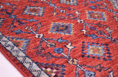 A close-up of a red patterned Afghan Chobi mat with blue, beige, and white geometric motifs and a decorative border.