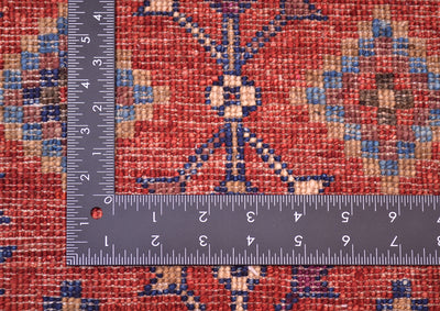 A close-up of the hand-knotted back of a patterned Afghan Chobi tribal design mat in red, blue, beige, and brown tones, shown with a metal ruler to indicate scale of the knotting.
