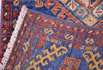 Close-up of the corner turned up to show the hand-knotted weave of a short Afghan Chobi runner rug showing a blue field with orange, yellow, and cream geometric motifs, white fringe, and part of a red border.