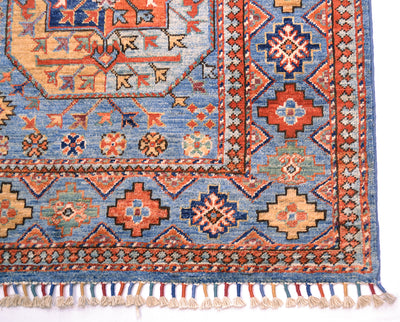 A close-up of a patterned Afghan area rug corner showing a blue field with orange, red, beige, and black geometric motifs and tassels along the bottom edge.