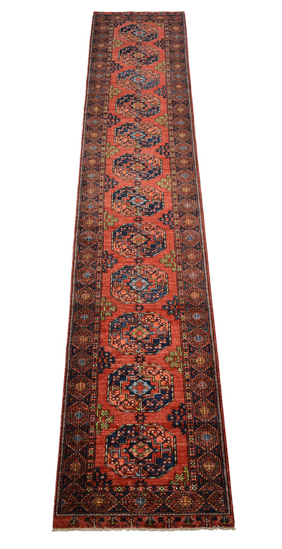 A 5m long runner rug with a traditional Afghan Ersari design, featuring a vibrant orange/rust field, dark navy blue medallions, and intricate geometric borders in beige and green.
