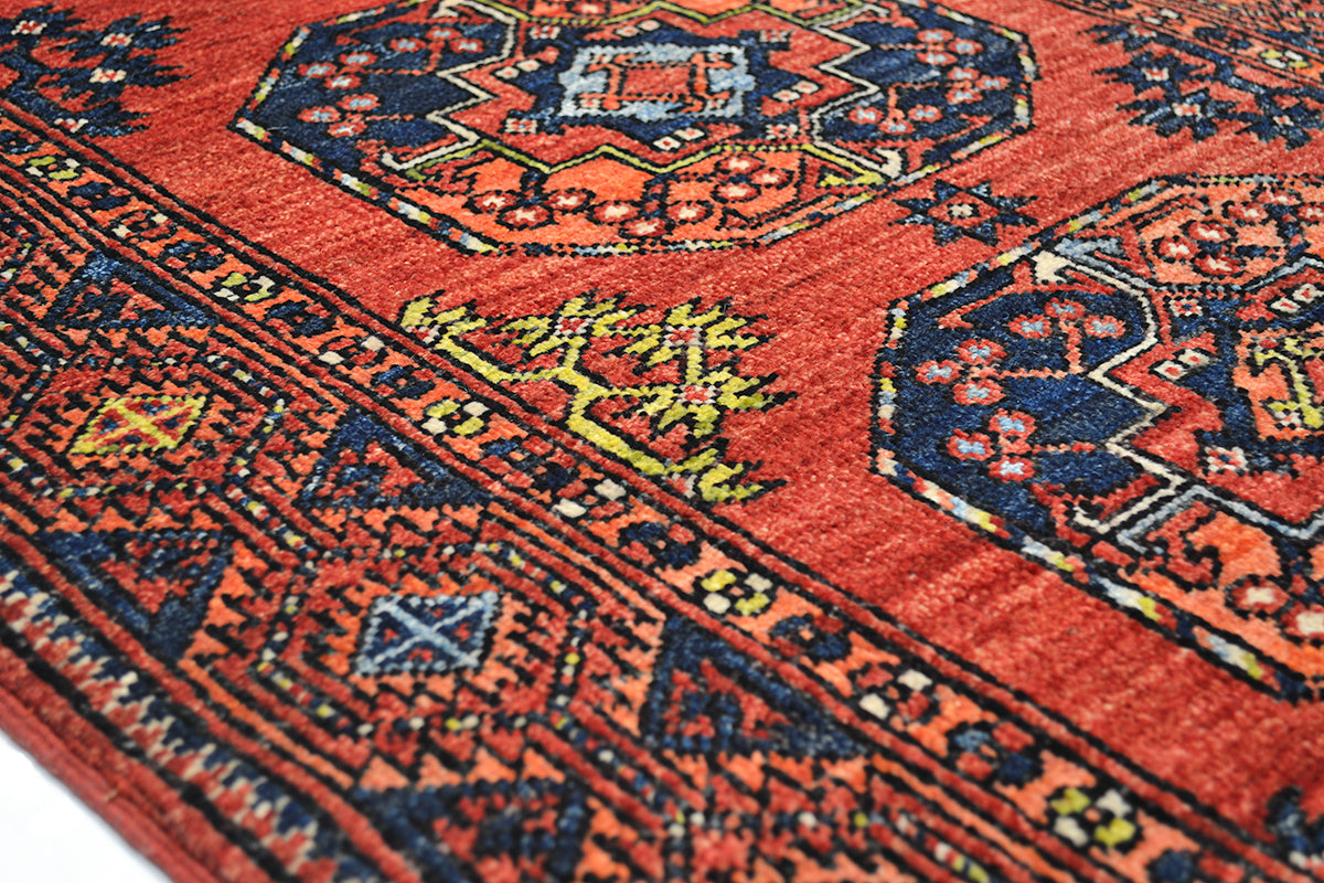A close-up of a traditional patterned Afghan long runner rug with a warm rust-red field, dark navy geometric borders, and small yellow and light blue floral accents.