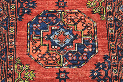 A close-up of a traditional handwoven Afghan runner rug featuring a vibrant red field with a central octagonal medallion in navy blue, orange, and light blue, surrounded by floral motifs and patterned borders.