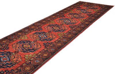 A long oriental Afghan corridor runner rug with a warm rust field, dark navy blue central medallions, and intricate multicolour floral patterns, shown diagonally on a white background.