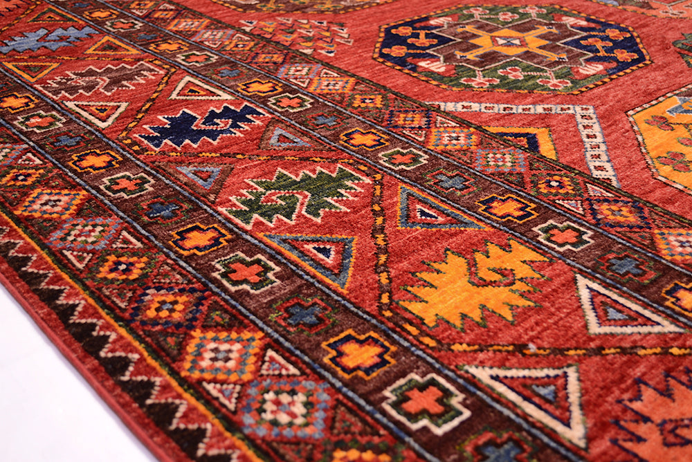 A close-up of a traditional Afghan Ersari carpet with a warm red-orange base and geometric motifs in brown, navy, yellow, green, and cream.