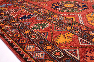 A close-up of a traditional Afghan Ersari carpet with a warm red-orange base and geometric motifs in brown, navy, yellow, green, and cream.