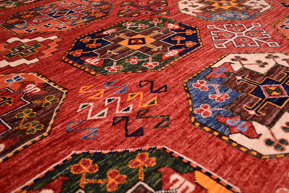 A close-up of a traditional large hand-knotted Ersari carpet with a dominant rust field and intricate geometric motifs in blue, green, yellow, and brown.