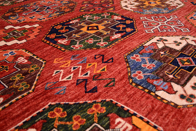 A close-up of a traditional large hand-knotted Ersari carpet with a dominant rust field and intricate geometric motifs in blue, green, yellow, and brown.