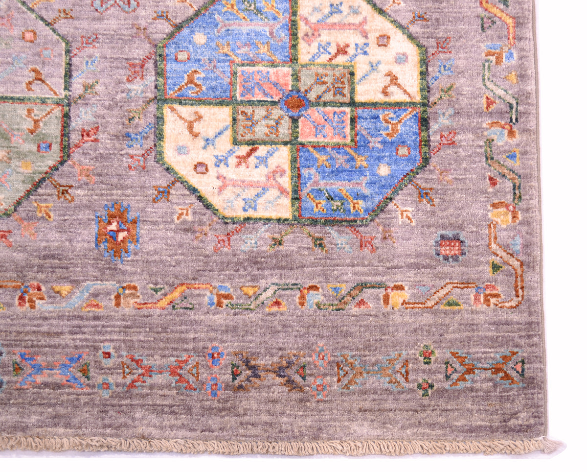 A close-up of a traditional hand-knotted rug showing its detailed pattern. The Afghan Fil-Pai rug features a muted grey field with a central octagonal medallion in blue, cream, and pink, surrounded by geometric borders and a fringed edge.