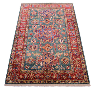 A rectangular traditional area Kazak rug with an intricate geometric and floral pattern, featuring a green centre field, red borders, and blue and beige accents, shown flat against a white background.
