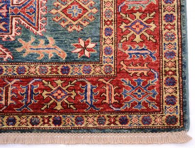 A close-up of a traditional patterned rug corner showing a red field with blue and beige geometric motifs, a dark blue-green border, and cream fringe.