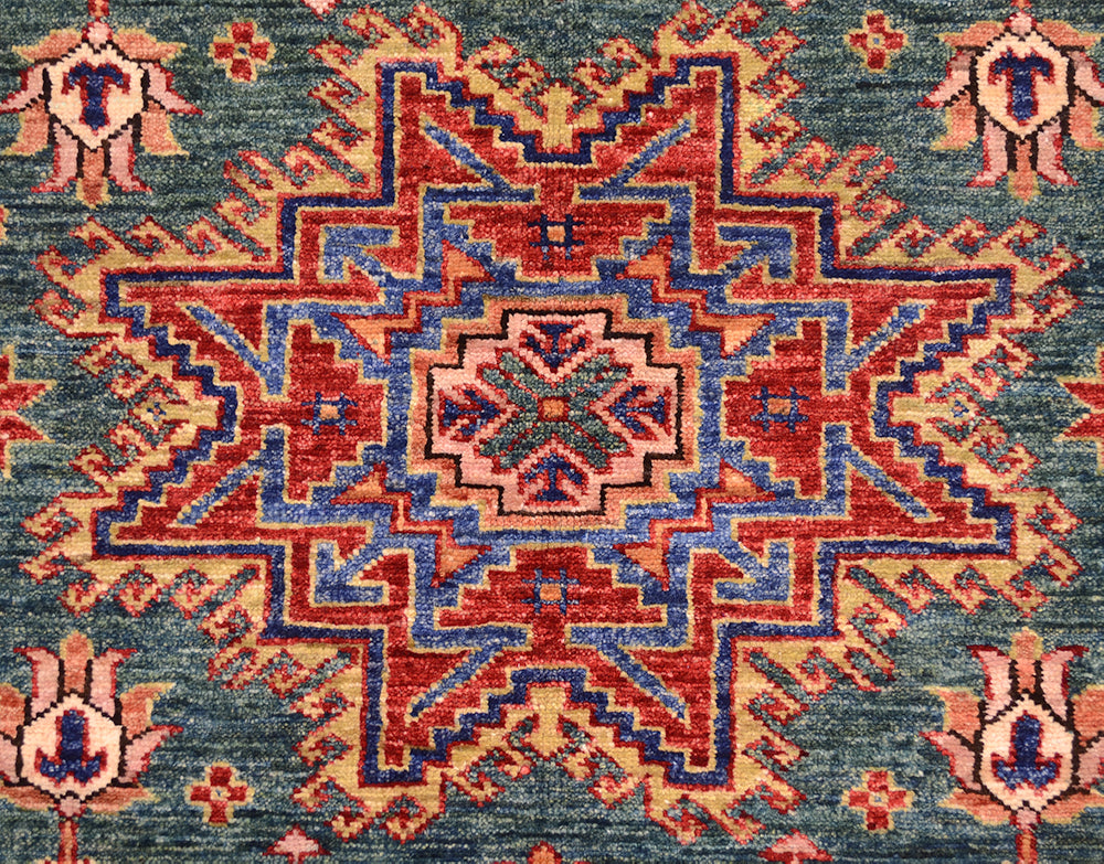 A close-up of a traditional patterned rug with a central medallion and corner motifs. The design features red, blue, and beige geometric and floral elements on a muted blue-gray background.