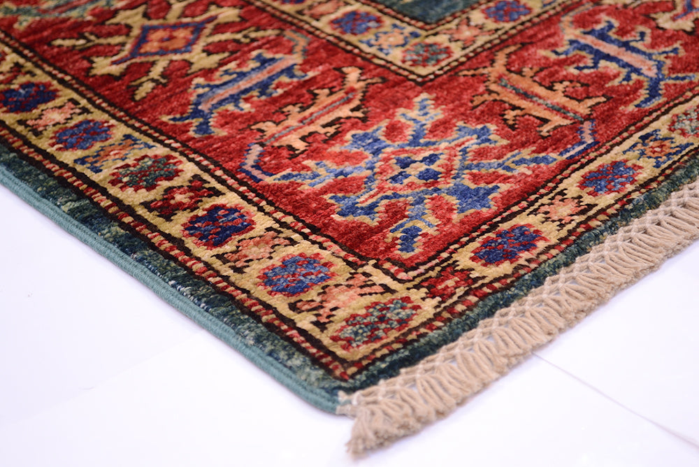 A close-up of a traditional patterned rug corner showing a red field with blue and beige floral motifs, a dark border, and cream fringe on a white floor.