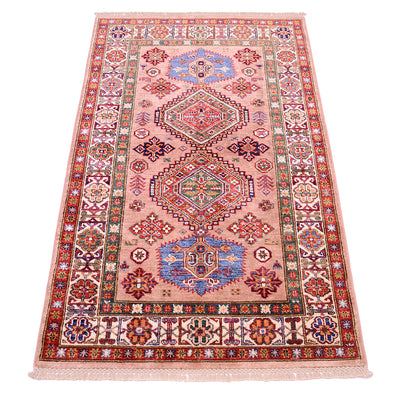 A rectangular fine handmade pink Kazak Rug with a complex geometric and floral pattern, featuring a dusty pink field, red borders, and blue and green medallions, shown flat against a white background.