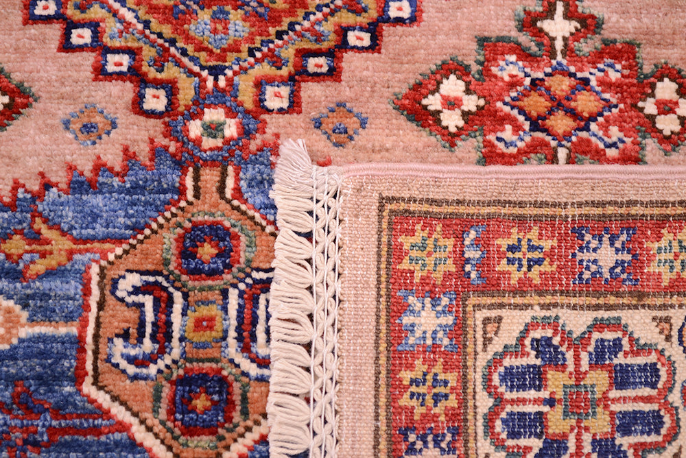 Close-up of the hand-knotted underside of a patterned Afghan Kazak rug corner showing cream fringe, with a pink field and red, blue, and beige geometric motifs.