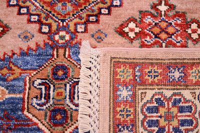 Close-up of the hand-knotted underside of a patterned Afghan Kazak rug corner showing cream fringe, with a pink field and red, blue, and beige geometric motifs.