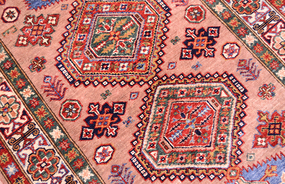 A close-up of a traditional Kazak handwoven rug with a dusty pink field, featuring large hexagonal medallions and geometric motifs in red, blue, green, and cream.