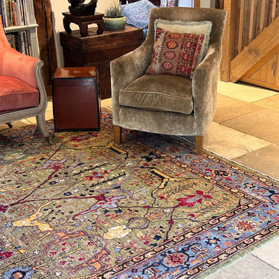 A large, ornate traditional-style soft handmade Green Garous Carpet with a complex multicolour pattern in blue, red, beige, and green, styled in a rustic living room setting with two armchairs and a wooden trunk.