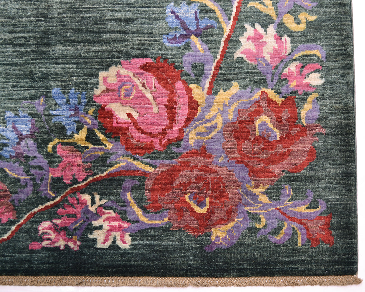 Close-up of a handwoven Afghan Karabagh carpet showing a floral pattern with large pink and red flowers, purple and blue foliage, and a dark green background. A narrow beige fringe is visible along the bottom edge.