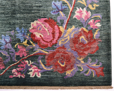 Close-up of a handwoven Afghan Karabagh carpet showing a floral pattern with large pink and red flowers, purple and blue foliage, and a dark green background. A narrow beige fringe is visible along the bottom edge.